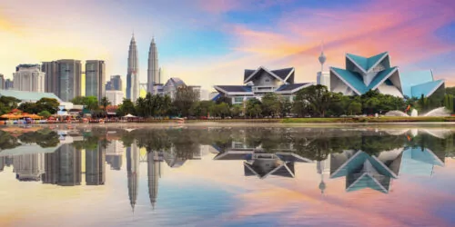 Malaysia skyline with Petronas Towers and colorful sunset reflecting in a calm lake.